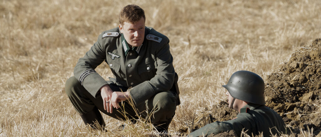 "World War II German officer engaged in conversation with a soldier in a foxhole, showcasing a moment of camaraderie amidst the chaos of war. The officer, in a standard Wehrmacht uniform and peaked cap, leans slightly towards the soldier, who is dressed in combat attire and equipped with a rifle. The foxhole is surrounded by earth and camouflage netting, providing a glimpse into the harsh realities of trench warfare. Their expressions convey a mix of seriousness and determination as they discuss strategy or share words of encouragement."
